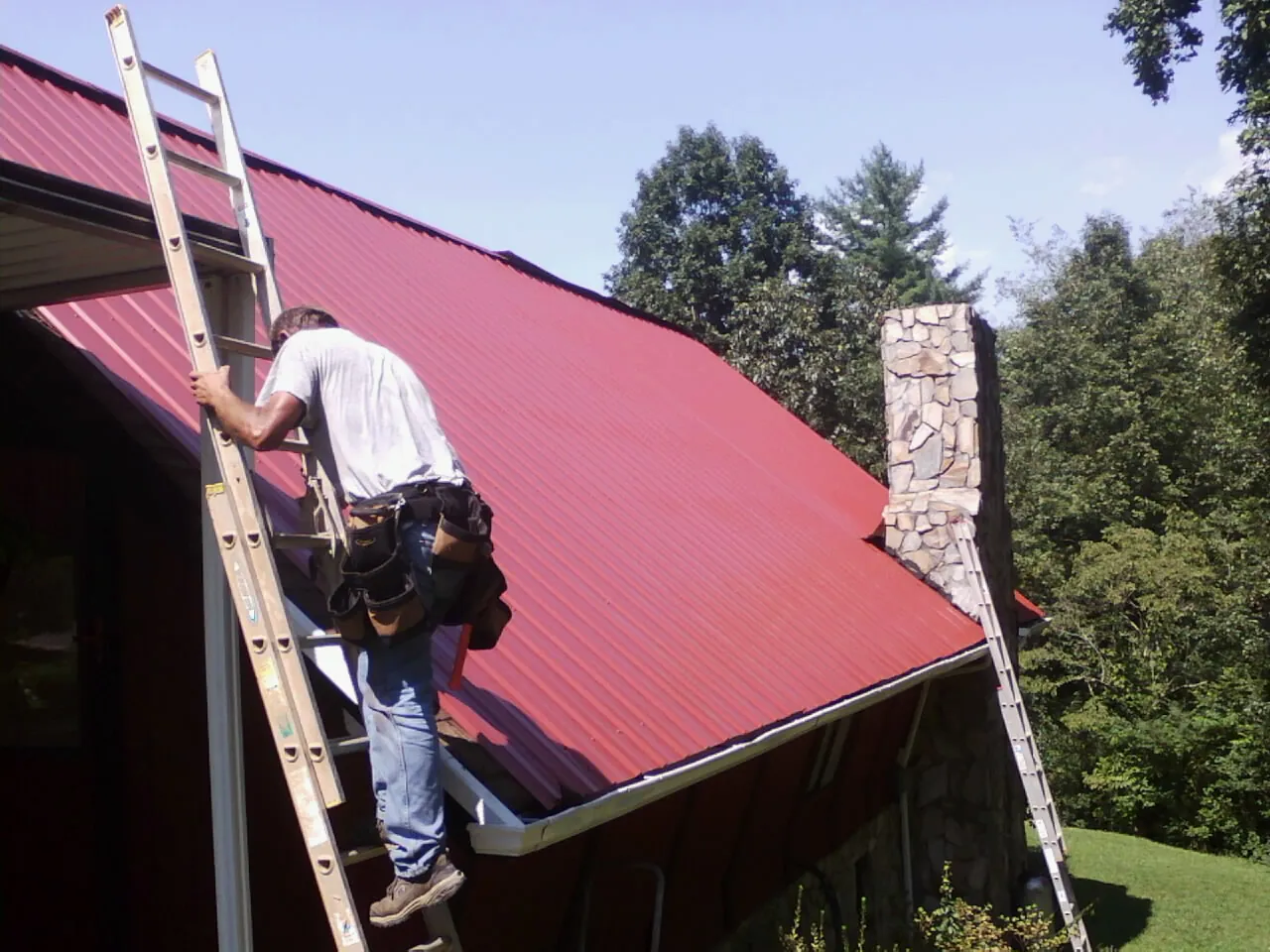 Roofer on a ladder beside a red metal roof and stone chimney — Soffit & Fascia Repair by Roofing Brothers in Nicholasville
